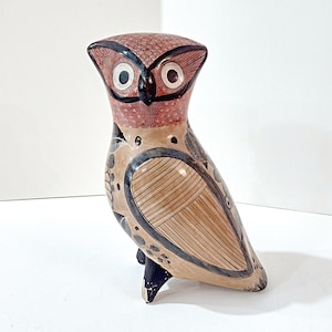 May include: A ceramic owl figurine with a detailed design. The owl has a reddish-brown head and a tan body with black and white accents. The eyes are large and white with black pupils. The owl is standing on a white surface.