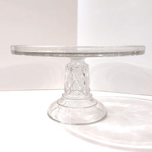 May include: A clear glass cake stand with a round top and a decorative, faceted pedestal base. The stand is elevated and sits on a white surface. The design is simple and elegant.
