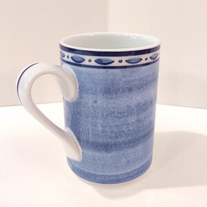 May include: A ceramic mug with a blue watercolor design. The mug has a white handle and a decorative blue band around the rim. The mug is approximately 4 inches tall.