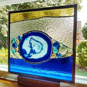 May include: Stained glass panel with a blue agate design, framed in black. The panel features a blue and white agate center, surrounded by blue, teal, and yellow glass beads. The top section is gold and silver, with a blue wave at the bottom.