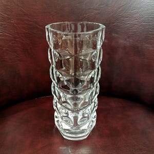 May include: A clear glass vase with a geometric design. The vase has a hexagonal base and a series of stacked, diamond-shaped patterns along its body. The top of the vase is also hexagonal. The vase is empty and sits on a dark red surface.