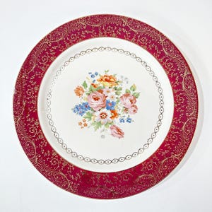 May include: A round ceramic dinner plate featuring a wide, deep magenta border decorated with intricate gold filigree patterns. The center of the plate is white and displays a colorful floral bouquet of pink, orange, and blue flowers.
