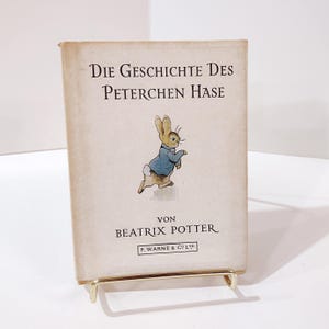 May include: A vintage book titled "Die Geschichte Des Peterchen Hase" by Beatrix Potter, featuring an illustration of a rabbit in a blue jacket. The book is displayed on a gold-colored stand.