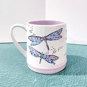 May include: White ceramic mug with a dragonfly design. The mug features two dragonflies with blue and purple floral wings. The interior and base of the mug are light pink. The handle is white.