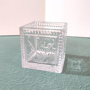 May include: A clear, square glass votive candle holder. The sides feature a decorative pattern with a floral design. The top rim has a series of small, raised rectangles. The candle holder sits on a light blue surface.