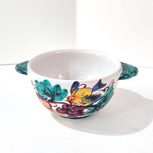 May include: White ceramic bowl with a hand-painted design featuring a yellow and blue bird, green leaves, and purple grapes. The bowl has two green handles and is suitable for serving soups or other dishes.