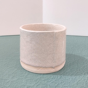 May include: A handmade, cylindrical ceramic cup with a speckled, light gray exterior and a cream-colored base. The cup has a smooth, rounded rim and a simple, minimalist design. Perfect for holding beverages or as a decorative piece.