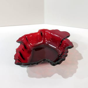 May include: A ruby red glass dish with a ruffled, leaf-like design. The dish has a glossy finish and a dark, almost black, rim. The bowl is sitting on a white surface.