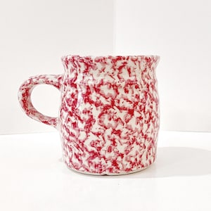 May include: A ceramic mug with a red and white speckled pattern. The mug has a rounded body, a curved handle, and a slightly flared rim. The speckled design covers the entire surface of the mug.