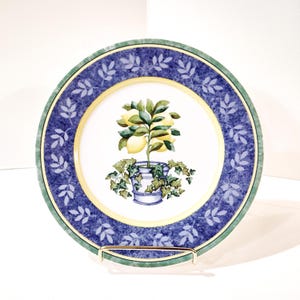 May include: A decorative ceramic plate featuring a lemon tree in a blue and white pot. The plate has a blue border with a leaf pattern, a yellow inner ring, and a green outer rim. The plate is displayed on a gold-colored metal stand.