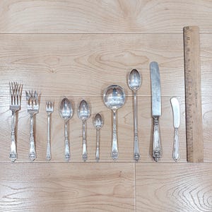 May include: A collection of antique silverware, including forks, spoons, a ladle, a knife, and a butter knife. The silverware is silver-colored with decorative detailing. A wooden ruler is placed next to the silverware for scale.