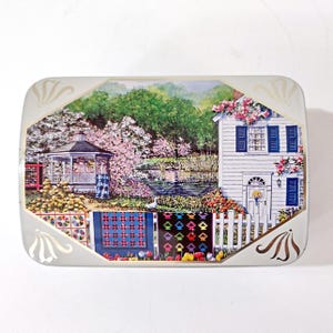 May include: A rectangular metal tin featuring a printed landscape scene of a white house with blue shutters, a gazebo, a pond, and a white picket fence. Several colorful patterned quilts are draped over the fence in the foreground.