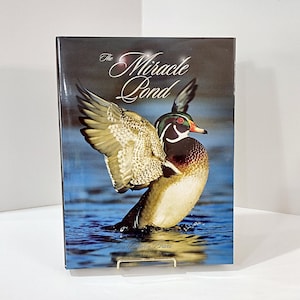 May include: A hardcover book titled "The Miracle Pond" featuring a vibrant image of a Wood Duck in flight over water. The duck has brown, white, and black feathers, with a red and white beak. The author's name, Middleton Evans, is printed at the bottom.