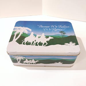 May include: A rectangular metal tin featuring a blue and green desert landscape with white silhouettes of figures riding camels under palm trees. The lid displays the text "Because We Believe, We Rejoice!" in a white script font against a blue sky.