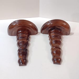 May include: Two wooden bottle stoppers with a mushroom-shaped top and a ridged stem. The stoppers are made of a dark brown wood with a glossy finish. The design is simple and elegant, suitable for wine bottles.