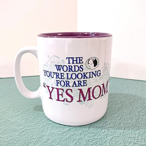 May include: A white ceramic mug with a purple interior and handle. The front features a blue and purple graphic with the text "THE WORDS YOU'RE LOOKING FOR ARE "YES MOM"" and a small circular icon of a dog silhouette in the upper right corner.