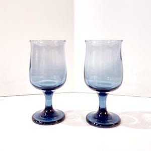 May include: Two blue-tinted wine glasses with a tulip-shaped bowl and a stem. The glasses have a dark blue base and stem, with a lighter blue hue towards the top. The glasses are set against a white background.