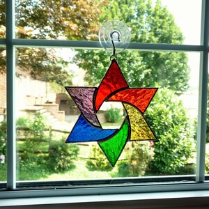 Pu&ograve; includere: Una stella di David in vetro colorato arcobaleno appesa a una finestra. La stella &egrave; composta da sei punte ed &egrave; circondata da un bordo in vetro trasparente.
