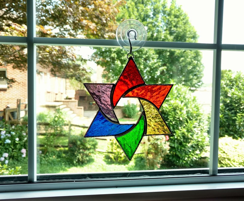 Pu&ograve; includere: Una stella di David in vetro colorato arcobaleno appesa a una finestra. La stella &egrave; composta da sei punte ed &egrave; circondata da un bordo in vetro trasparente.