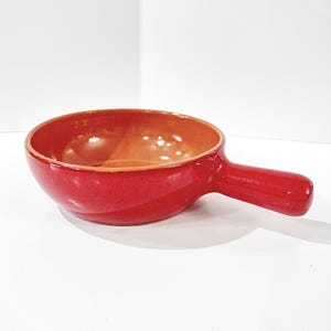 May include: A red ceramic serving bowl with a handle. The bowl has a rounded shape with a smooth, glossy finish. The interior is a lighter shade of orange-brown. The handle is a solid, cylindrical shape, making it easy to hold.
