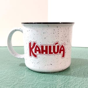 May include: White speckled ceramic mug with the red "Kahlúa" logo. The mug has a black rim and a white handle. The mug is sitting on a light green surface.