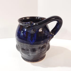 May include: Handmade ceramic mug with a dark blue and gray glaze. The mug features a rounded body with a textured lower half and a large, curved handle. The interior is a deep blue, and the handle is a glossy black.