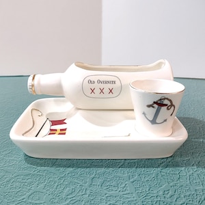 May include: A white ceramic nautical-themed set featuring a bottle-shaped container with the text "Old Overnite" and a shot glass with an anchor design. The set includes a rectangular tray with nautical flag details.