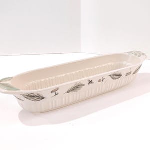 May include: An elongated, oval-shaped ceramic serving dish in an off-white color. The exterior features a vertical ribbed texture and a decorative pattern of dark green leaves, small bees, and tiny floral sprigs. It has integrated handles.