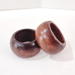 May include: Two dark brown wooden napkin rings. Each ring is a rounded cylinder shape with a smooth, polished finish. The rings are made of a dark wood, and the grain is visible. The rings are on a white surface.