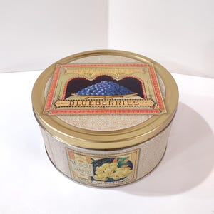 May include: A round, vintage tin with a gold lid and a beige body. The lid features a label with the word "BLUEBERRIES" and an illustration of blueberries. The body has a label that says "MUSCAT GRAPES".