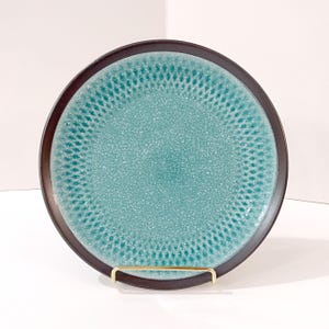 May include: A round ceramic plate featuring a speckled turquoise center with a darker teal geometric pattern radiating outward. The plate has a smooth, dark brown rim and is displayed upright on a thin, gold-toned metal wire stand.