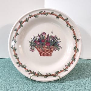 May include: A white ceramic plate with a floral basket design. The plate features a brown vine border with green leaves and a central image of a woven basket filled with pink, purple, and red flowers. A small insect is flying above the basket.