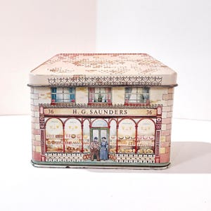 May include: A vintage tin shaped like a bakery shop, featuring a detailed facade with windows, a shop front, and the text "H.G. Saunders." The tin has a roof design and a decorative border. The shop front displays baked goods.