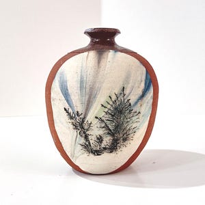 May include: A handcrafted ceramic vase with a unique design. The vase features a brown rim and top, with a white body adorned with blue and green brushstrokes and a black tree illustration. The vase is approximately 5 inches tall.