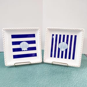May include: Two square white ceramic plates with a nautical theme. One plate has blue horizontal stripes, the other has vertical stripes. Each plate features a white seashell in the center and a rope-like border.