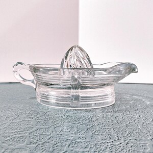 May include: Clear glass citrus juicer with a reamer and a pouring spout. The juicer has a handle and a ribbed design. The reamer is cone-shaped and has a textured surface. The juicer is designed for squeezing citrus fruits.