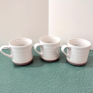 May include: Three white ceramic mugs with a horizontal ridged design and a mauve-colored base. Each mug has a curved handle. The mugs are arranged on a textured, teal-colored surface.