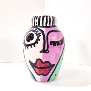 May include: A hand-painted ceramic vase with a whimsical face design. The vase features a black and white striped rim, a half-face design with a winking eye, and a red lip. The color palette includes pink, purple, and teal accents.