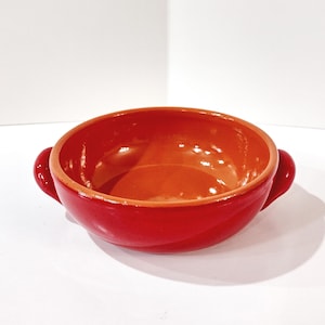 May include: A red ceramic bowl with two small handles. The interior is a lighter shade of red, and the exterior is a glossy, darker red. The bowl has a rounded shape and is likely used for serving food.
