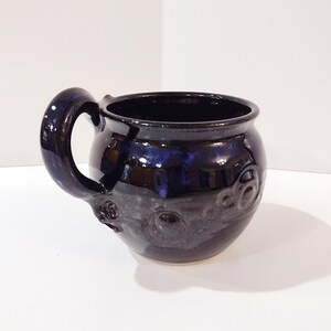 May include: A handmade, dark blue ceramic mug with a rounded body and a large, curved handle. The mug has a glossy finish with subtle blue highlights and embossed circular designs around the base.