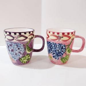 May include: Two ceramic mugs with floral designs. One mug has a purple and blue hydrangea pattern, while the other has a pink and blue hydrangea pattern. Both mugs have a decorative band at the top and a purple handle.