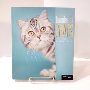 May include: A hardcover book titled "Gueules de CHATS" featuring a close-up of a gray and white cat with yellow eyes. The book has a light blue cover and is propped up by a gold-colored stand.