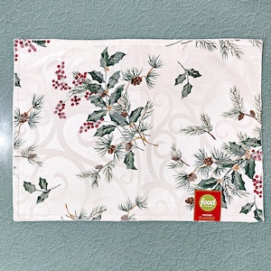May include: A rectangular fabric placemat featuring a cream-colored background with a subtle tonal swirl pattern. The design includes printed illustrations of green holly leaves, red berries, and brown pinecones. A red tag reads Food Network Holiday Placemat.