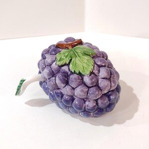 May include: A ceramic grape-shaped container with a lid. The container is a deep purple color, with a brown stem and a green leaf on top. The handle is white with green accents. The container is likely used for decorative purposes.