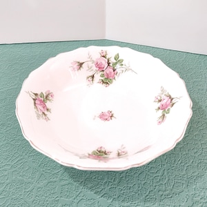 May include: A white, scalloped-edge serving bowl with a pink floral rose pattern. The bowl has a delicate gold trim and is set against a teal textured surface. The roses are in full bloom with green leaves.
