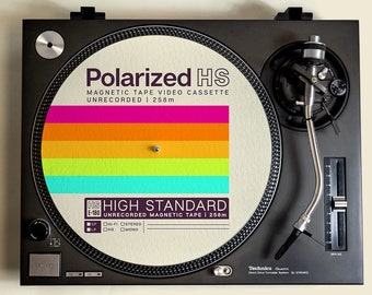 May include: A black turntable with a white slipmat featuring a colorful design and the text "Polarized HS Magnetic Tape Video Cassette Unrecorded | 258m" and "HS High Standard Unrecorded Magnetic Tape | 258m".