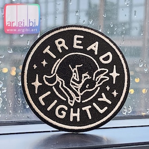 Peut inclure: Patch brodé noir avec des points de couture blancs. Le patch est rond et présente un animal endormi avec des étoiles et le texte "TREAD LIGHTLY".