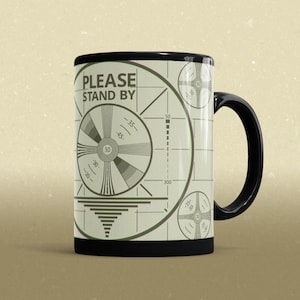 May include: Black coffee mug with a white design of a vintage television test pattern. The pattern includes the text "PLEASE STAND BY".