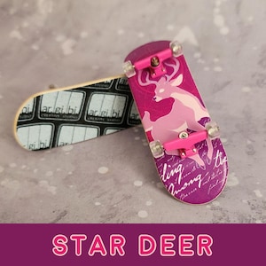May include: A pink fingerboard with a pink deer graphic and the text "wandering among the stars" on the bottom. The fingerboard has pink trucks and black wheels. The fingerboard is on a grey surface.