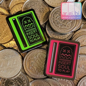 Può includere: Due spille in smalto nero e verde con un tema di videogiochi retrò. Le spille presentano un fantasma pixelato con il testo "Insert Soul To Play".